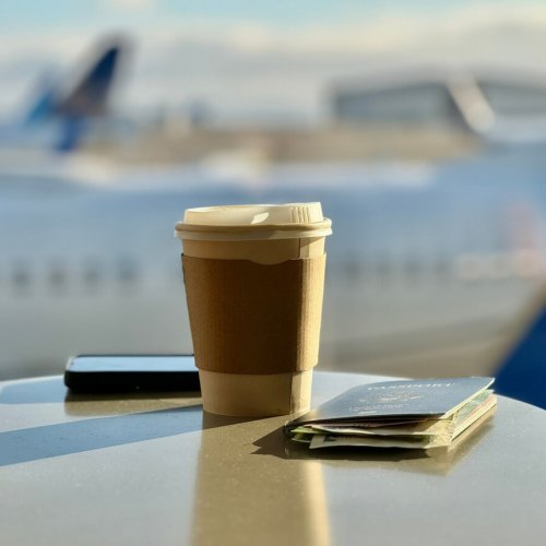 Early morning at an airport. A disposable cup and hot coffee sit on a table with passport and cell phone. Each object reflects off the table. Long shadows are cast from the early morning sun.